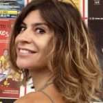 A woman with shoulder-length, wavy brown hair and a light smile looks over her shoulder. She is standing in front of a colorful wall covered with posters, radiating calm confidence like a reiki master skilled in energy healing. Reiki energy healing by jennybee alchemy
