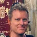 A smiling man with short blond hair stands indoors in front of a decorative maroon tapestry with gold symbols and hebrew writing, embodying the calm presence of a reiki master. Reiki energy healing by jennybee alchemy